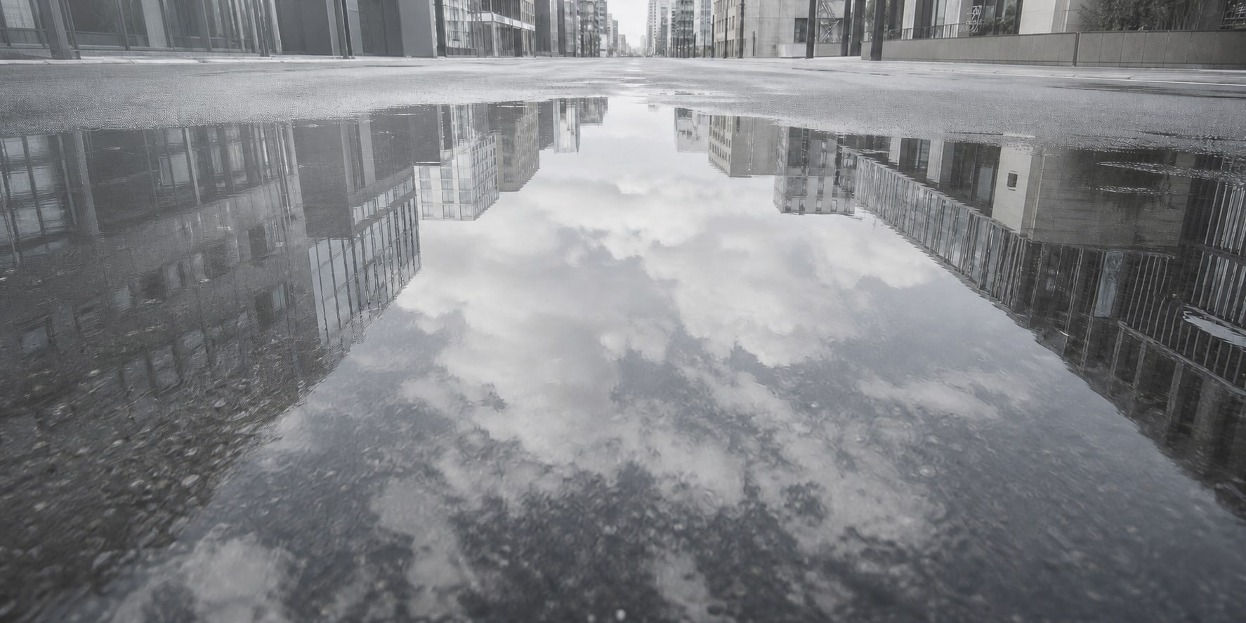 都市のビルに囲まれた濡れた道路の水たまりに空と雲が映る風景